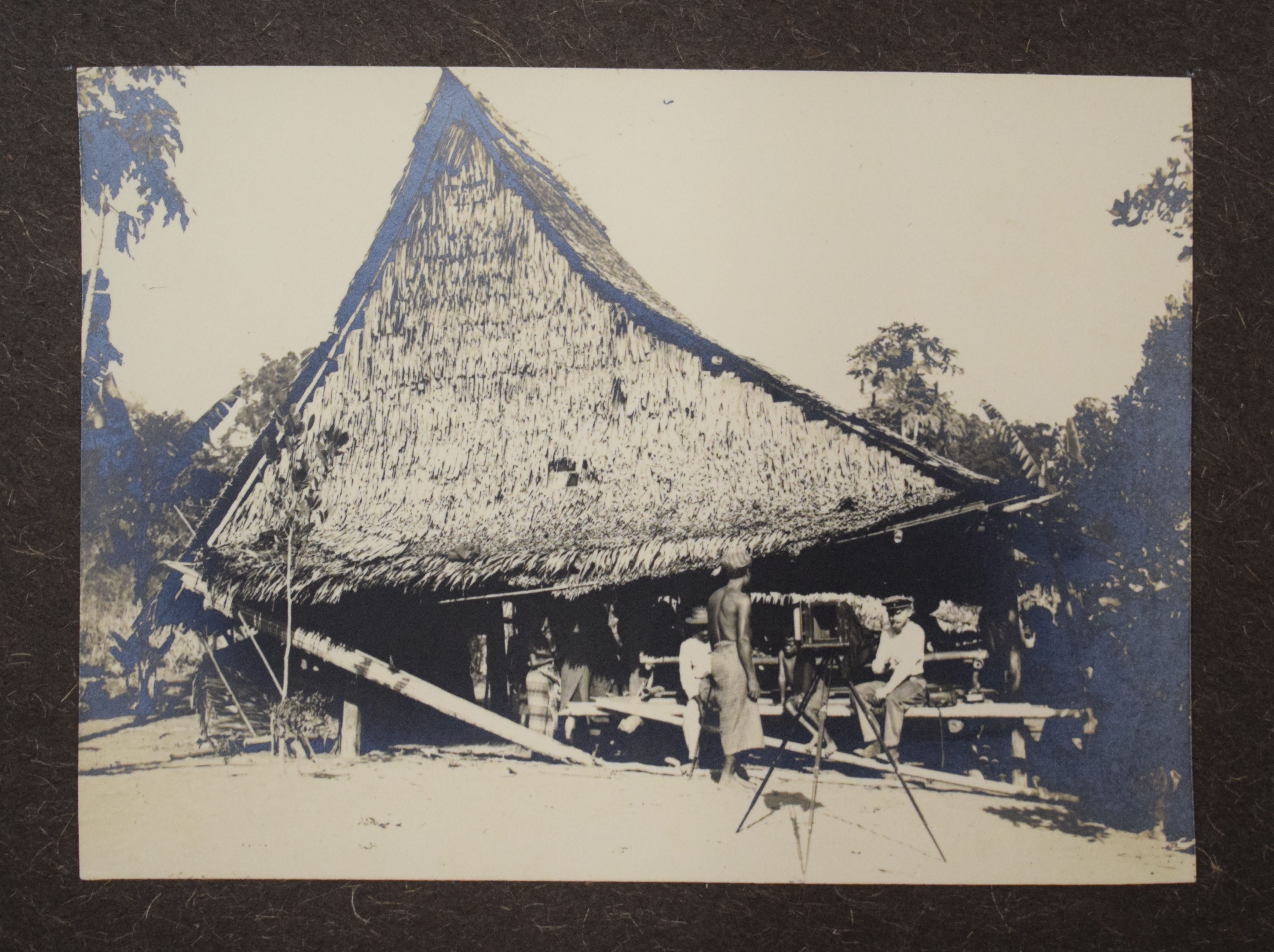 Article Image: This image was taken during the expedition, most probably by Erwin Stresemann. It shows several people in front of a wooden house, including Karl Deninger on the right, and a camera set up in front of them. This image is part of the collection at the Ethnologisches Museum, Berlin. Photo: L. Marten.