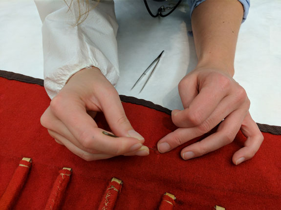 Article Image: Hands of a European trained museum conservator working on stabilizing a fabric, after negotiations with the knowledge holder of the source community concluded that the treatment was necessary to honor the object. Photo: Diana Gabler, 2017.